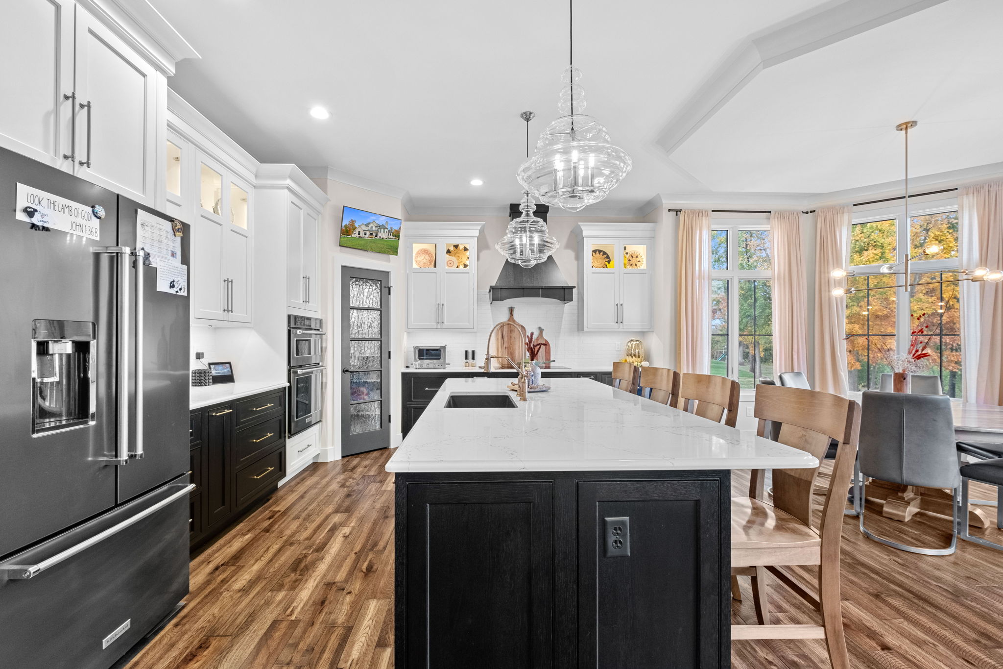 A modern kitchen featuring a large bay window, a drop ceiling, a spacious island with Cambria quartz countertops, and custom Shiloh cabinets. A bay window complements the dining room, enhanced by an elegant drop ceiling.