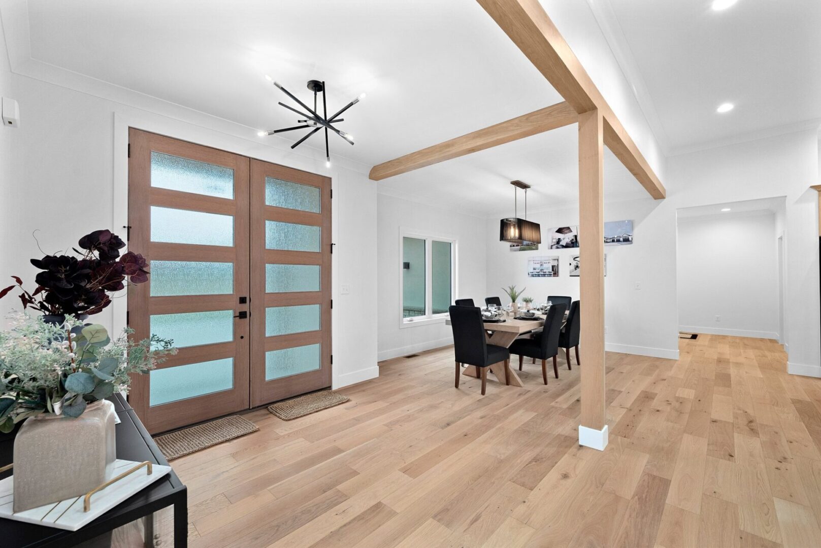 A grand entrance through contemporary modern double doors opens to a dining room embellished with modern lighting, white oak beams, and engineered hardwood floors.
