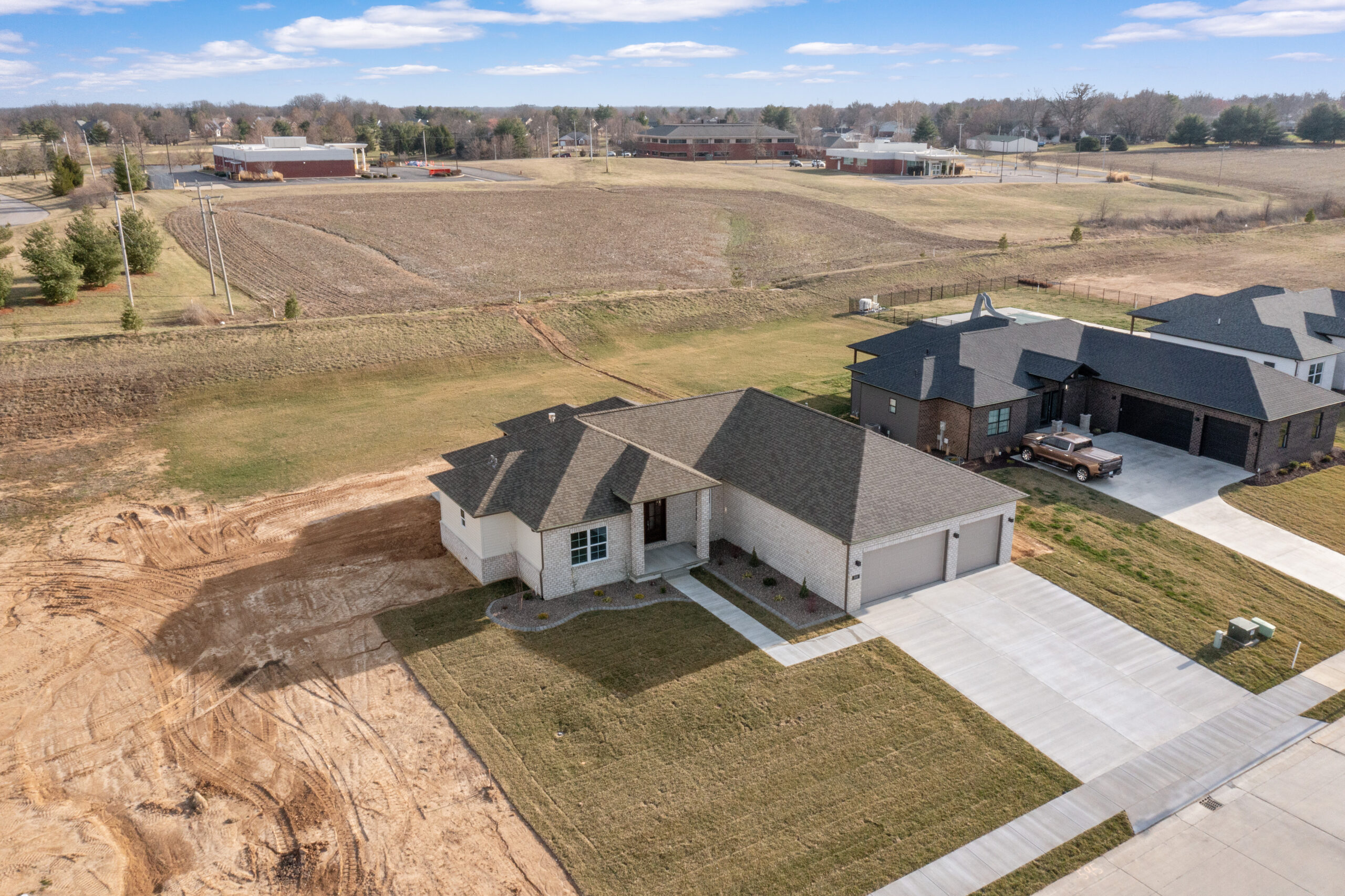 Drone photo of modern Legacy home. Legacy home designs featuring front and rear elevations with light toned brick exterior, creamy siding, and a stone pillar entryway. Three car garage, with 2 separate bays.