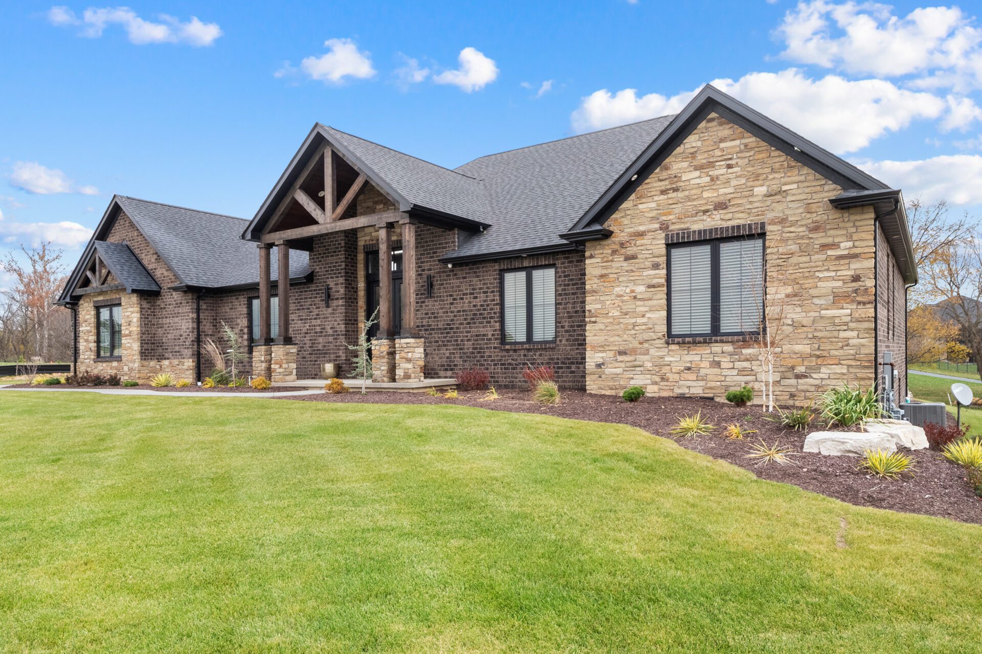 A contemporary mountain-style home features a stone and brick exterior. Over the front entrance, dark wooden beams stretch out, adding a touch of grandeur.