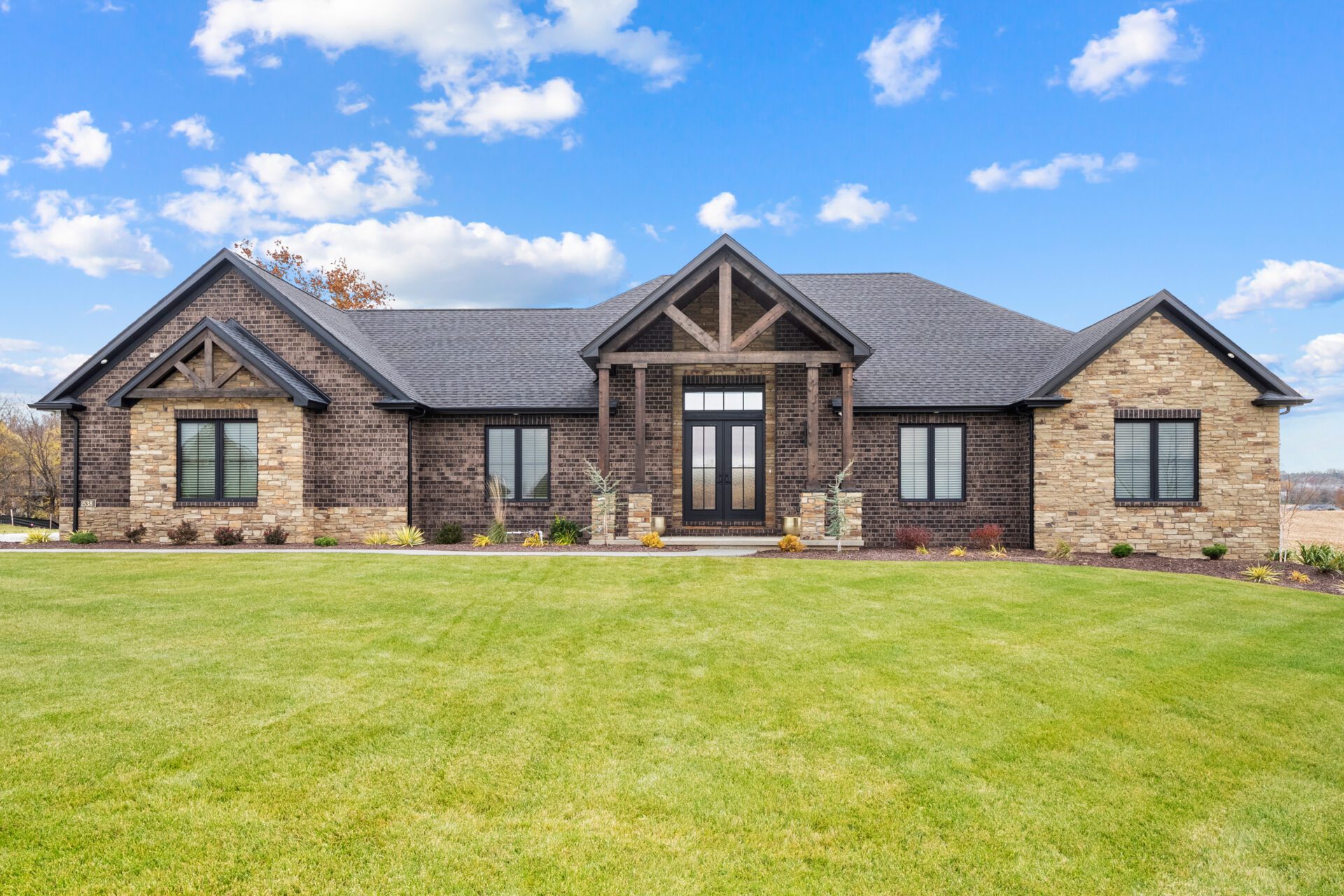 A contemporary mountain-style home features a stone and brick exterior. Over the front entrance, dark wooden beams stretch out, adding a touch of grandeur.