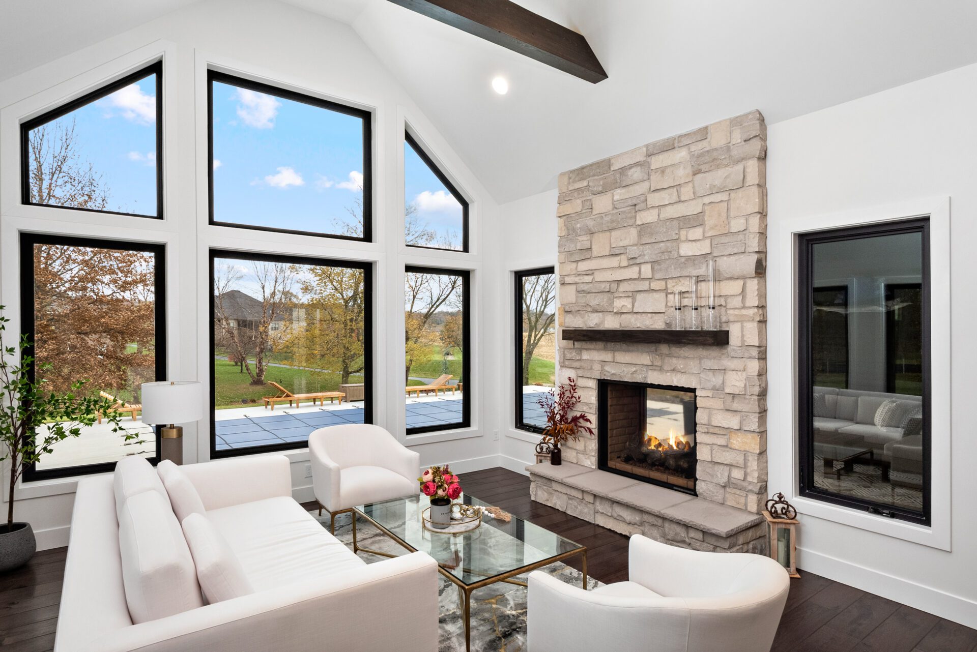 The heart room boasts a wall of windows offering an impeccable view of the pool. It showcases a double-sided, see-through fireplace that extends to the covered porch. A vaulted ceiling adorned with dark wooden beams stretches from the heart room into the kitchen.