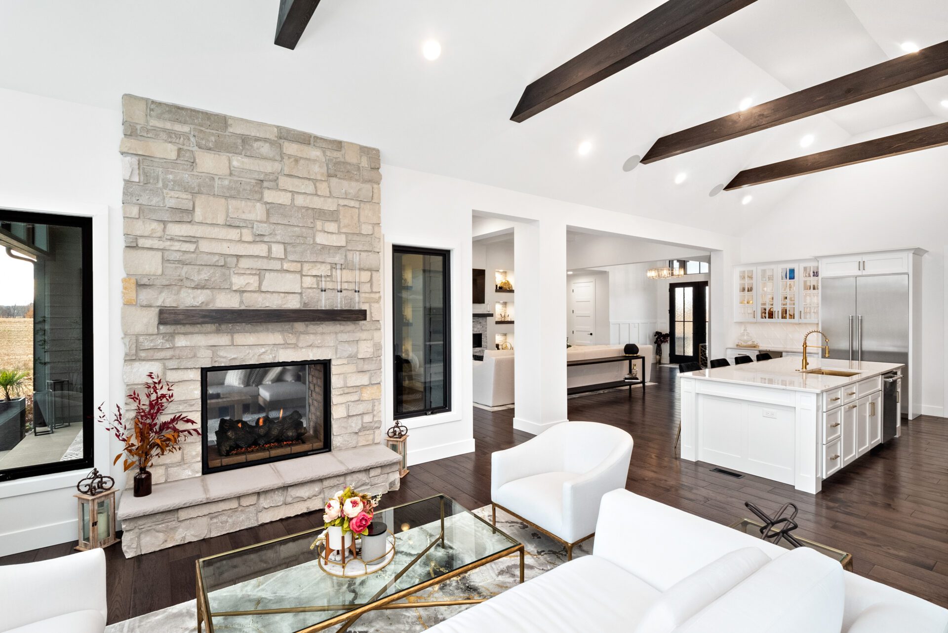 The heart room boasts a wall of windows offering an impeccable view of the pool. It showcases a double-sided, see-through fireplace that extends to the covered porch. A vaulted ceiling adorned with dark wooden beams stretches from the heart room into the kitchen.